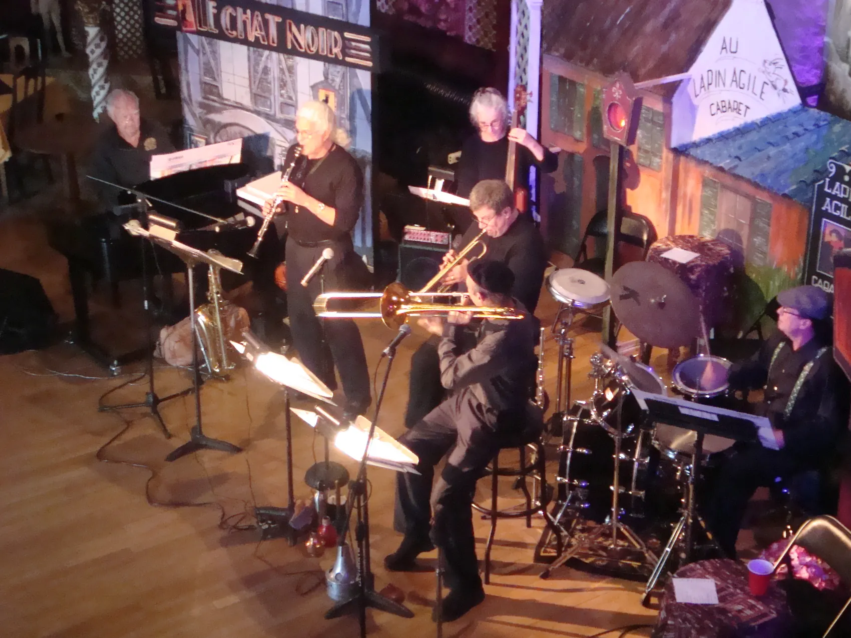 A wide view of a band playing before the Le Chat Noir cabaret backdrop.