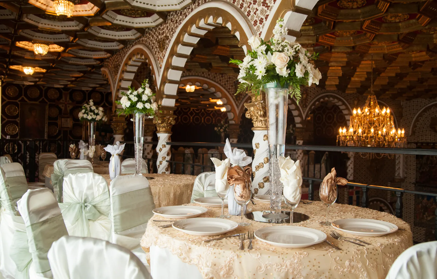 A head table with a tall floral centerpiece and a chandelier overhead.
