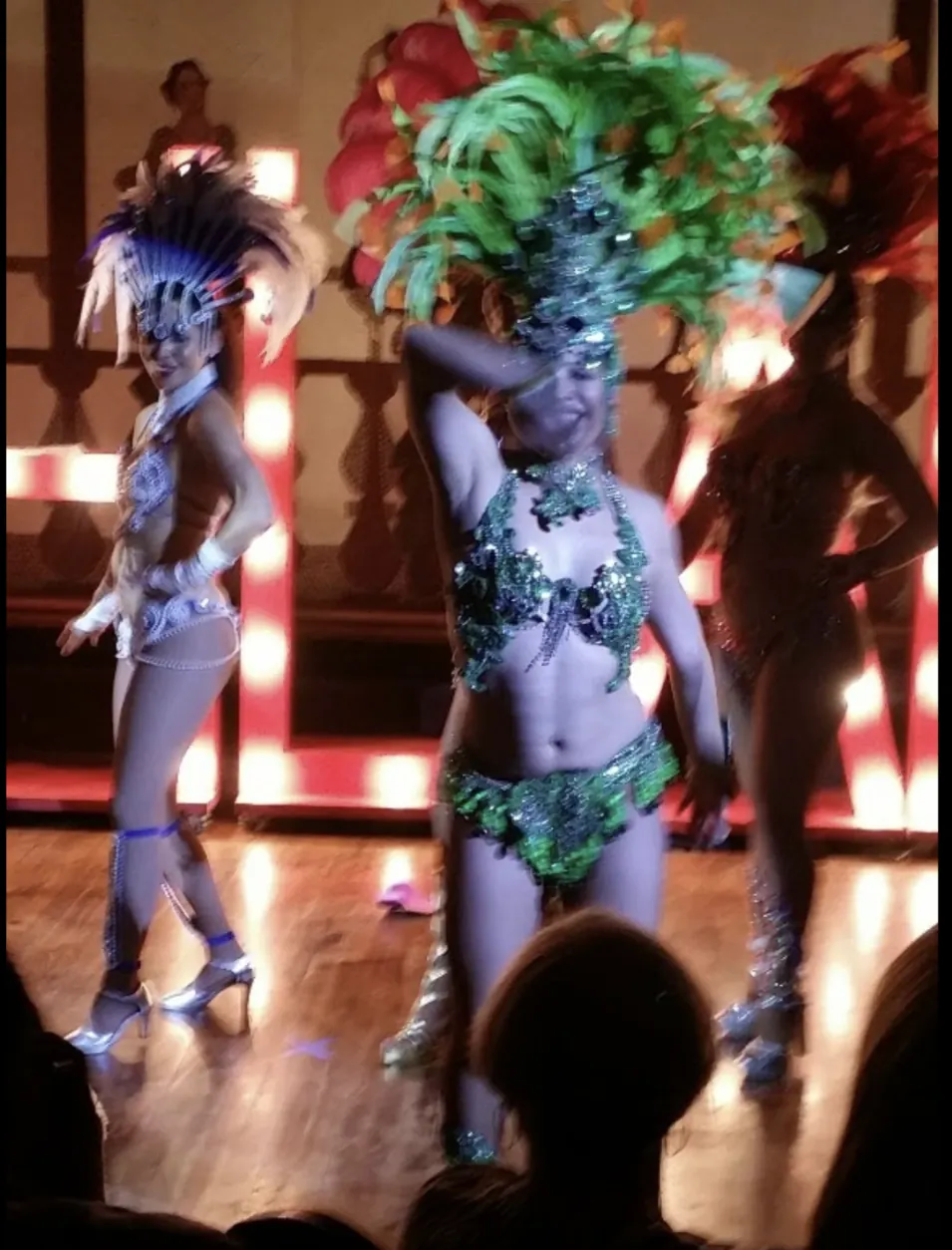 A dancer in a green-feathered showgirl costume performing on the lit floor.